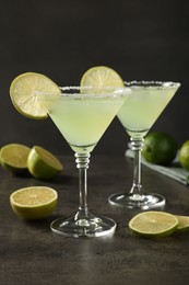 Delicious Margarita cocktail with ice cubes in glasses and limes on grey table