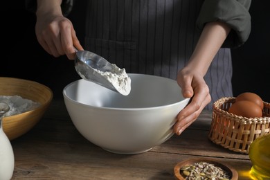 Making bread. Woman adding flour into bowl at wooden table on dark background, closeup