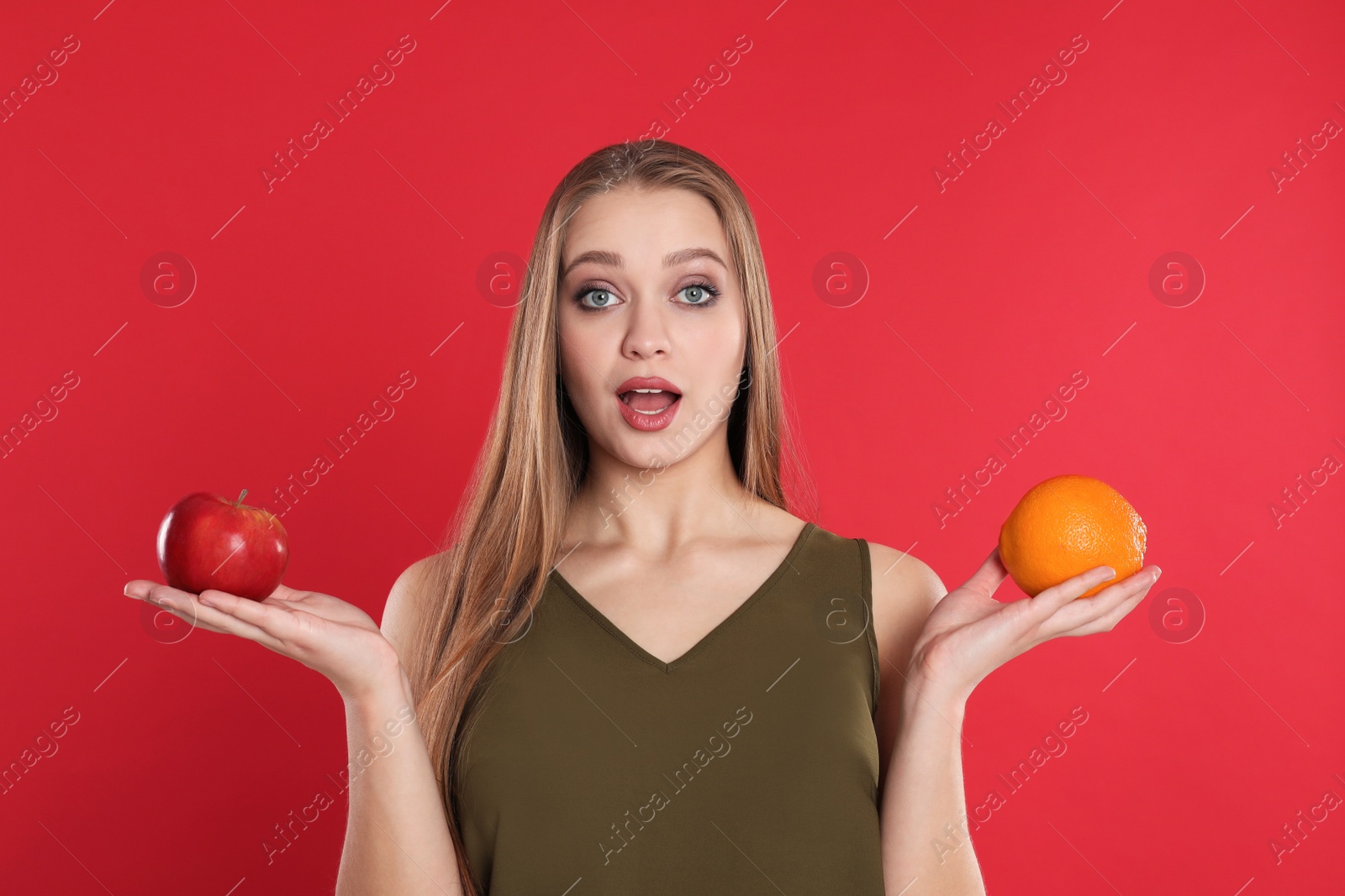 Photo of Emotional young woman with apple and orange on red background. Vitamin rich food