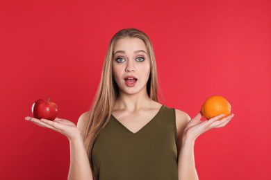 Photo of Emotional young woman with apple and orange on red background. Vitamin rich food