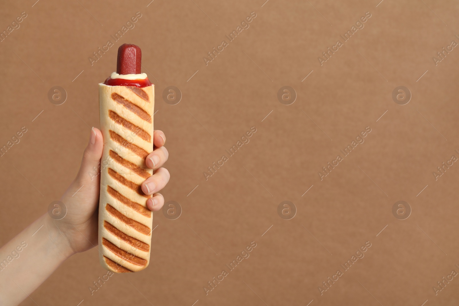 Photo of Woman holding delicious french hot dog on brown background, closeup. Space for text