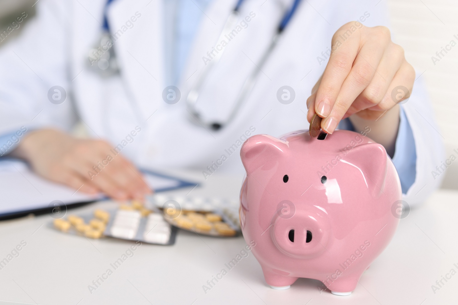 Photo of Doctor putting coin into piggy bank at white table, closeup