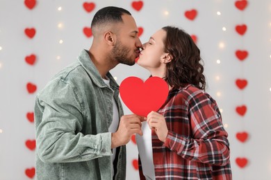 Photo of Lovely couple with red paper heart kissing indoors. Valentine's day celebration