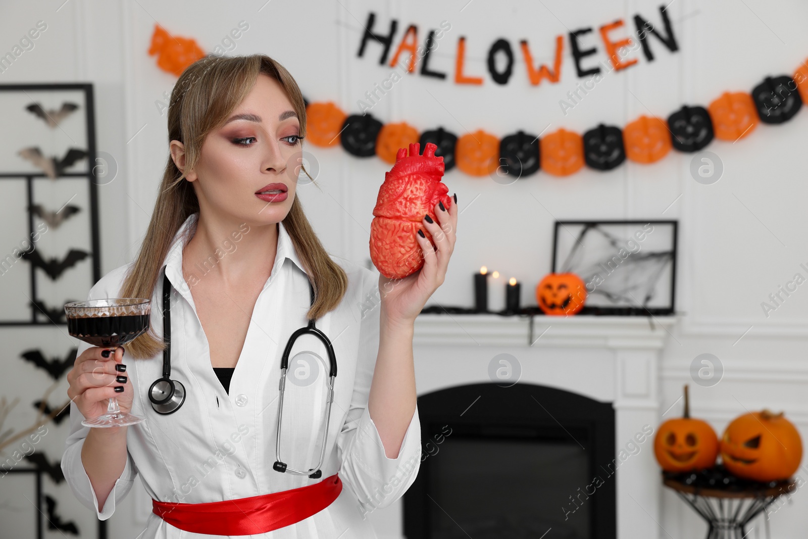 Photo of Woman in scary nurse costume with glass of wine and heart model indoors, space for text. Halloween celebration