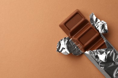 Photo of Delicious chocolate bar wrapped in foil on light brown background, top view. Space for text