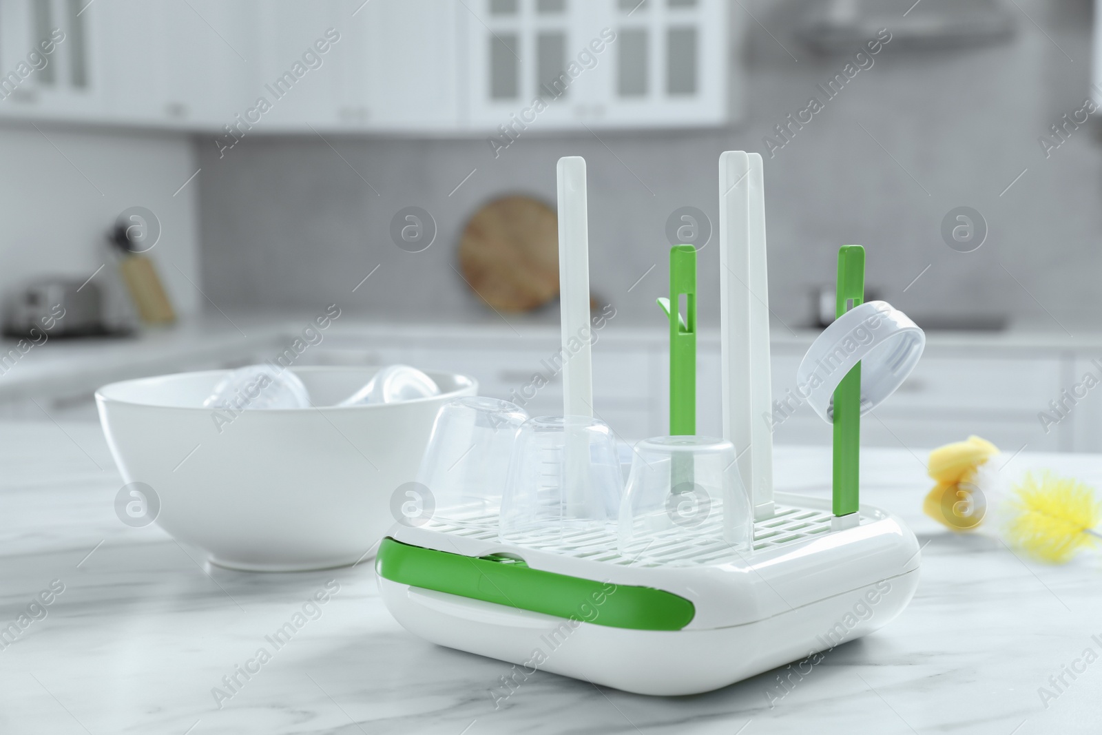 Photo of Dryer and bowl with baby bottle caps on white marble table in kitchen