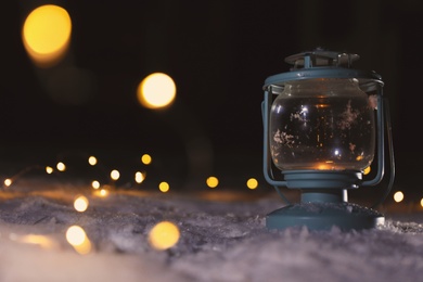 Photo of Lantern with burning candle and Christmas lights on white snow outdoors. Space for text