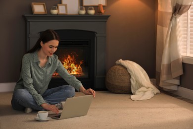 Photo of Beautiful young woman with laptop and cup of hot drink on floor near fireplace at home. Space for text