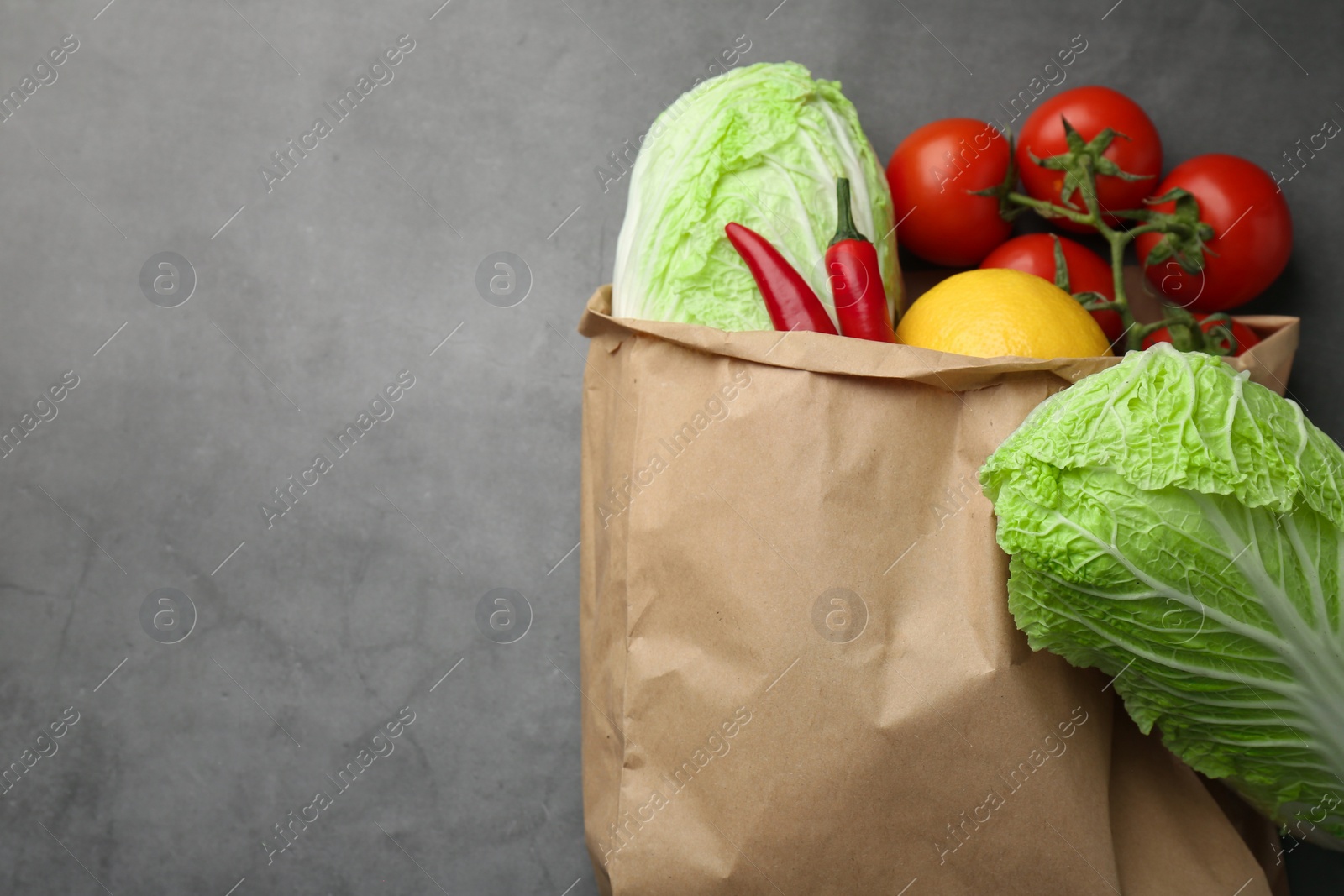 Photo of Paper bag with fresh Chinese cabbages, lemon, tomatoes and chili pepper on grey textured table, top view. Space for text