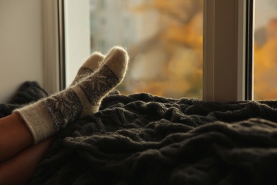 Photo of Woman in knitted socks relaxing on plaid near window at home, closeup