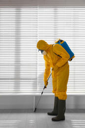 Photo of Male worker in protective suit spraying insecticide on wooden floor indoors. Pest control