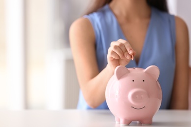 Photo of Woman putting coin into piggy bank at table indoors, closeup. Space for text