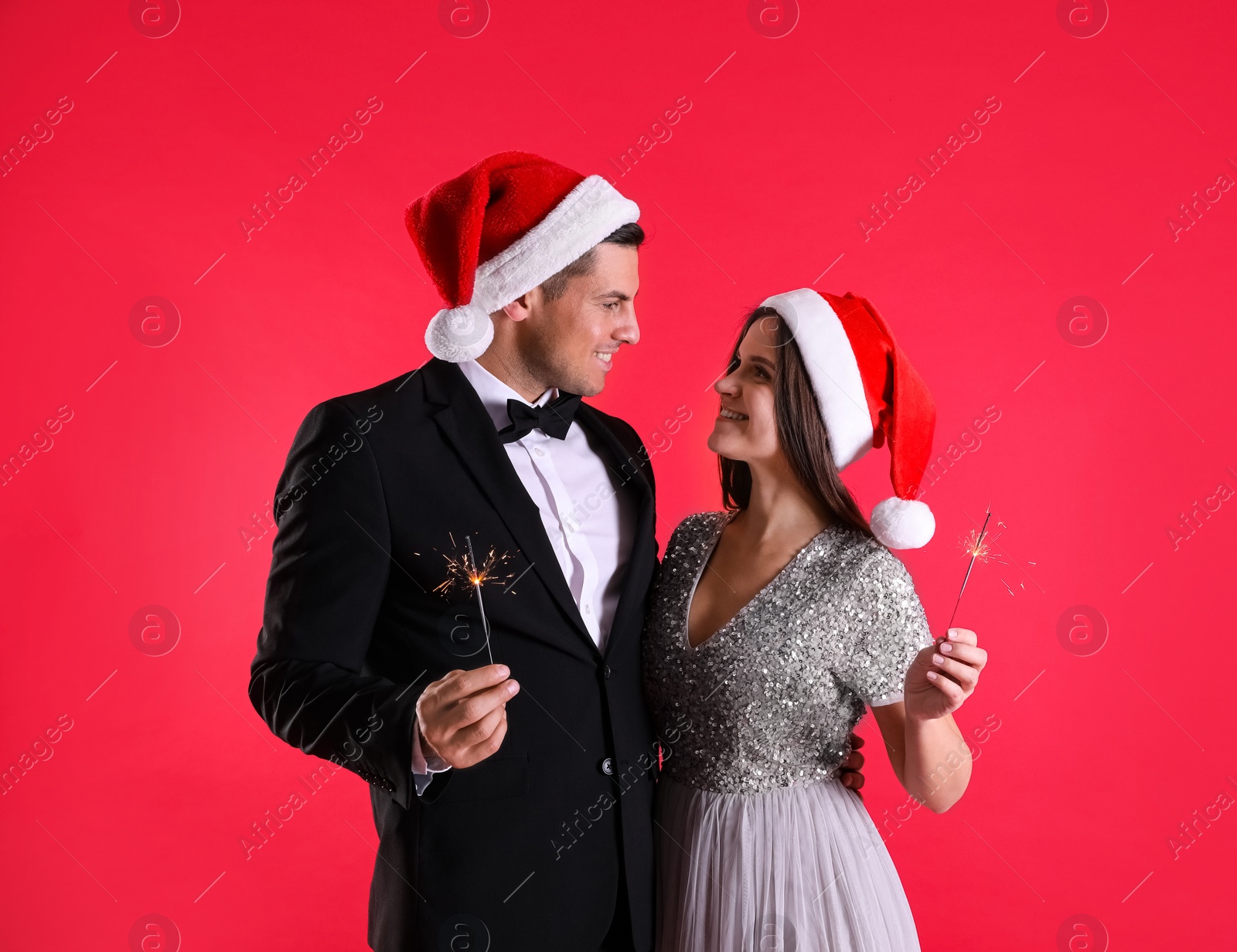 Photo of Beautiful happy couple in Santa hats with Christmas sparklers on red background