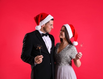 Photo of Beautiful happy couple in Santa hats with Christmas sparklers on red background