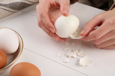 Woman peeling boiled egg at white wooden table, closeup