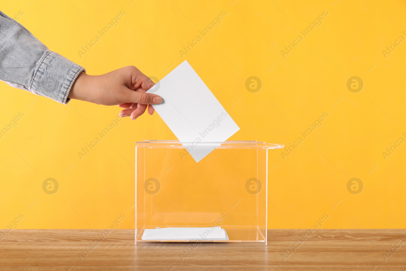 Photo of Woman putting her vote into ballot box on wooden table against orange background, closeup