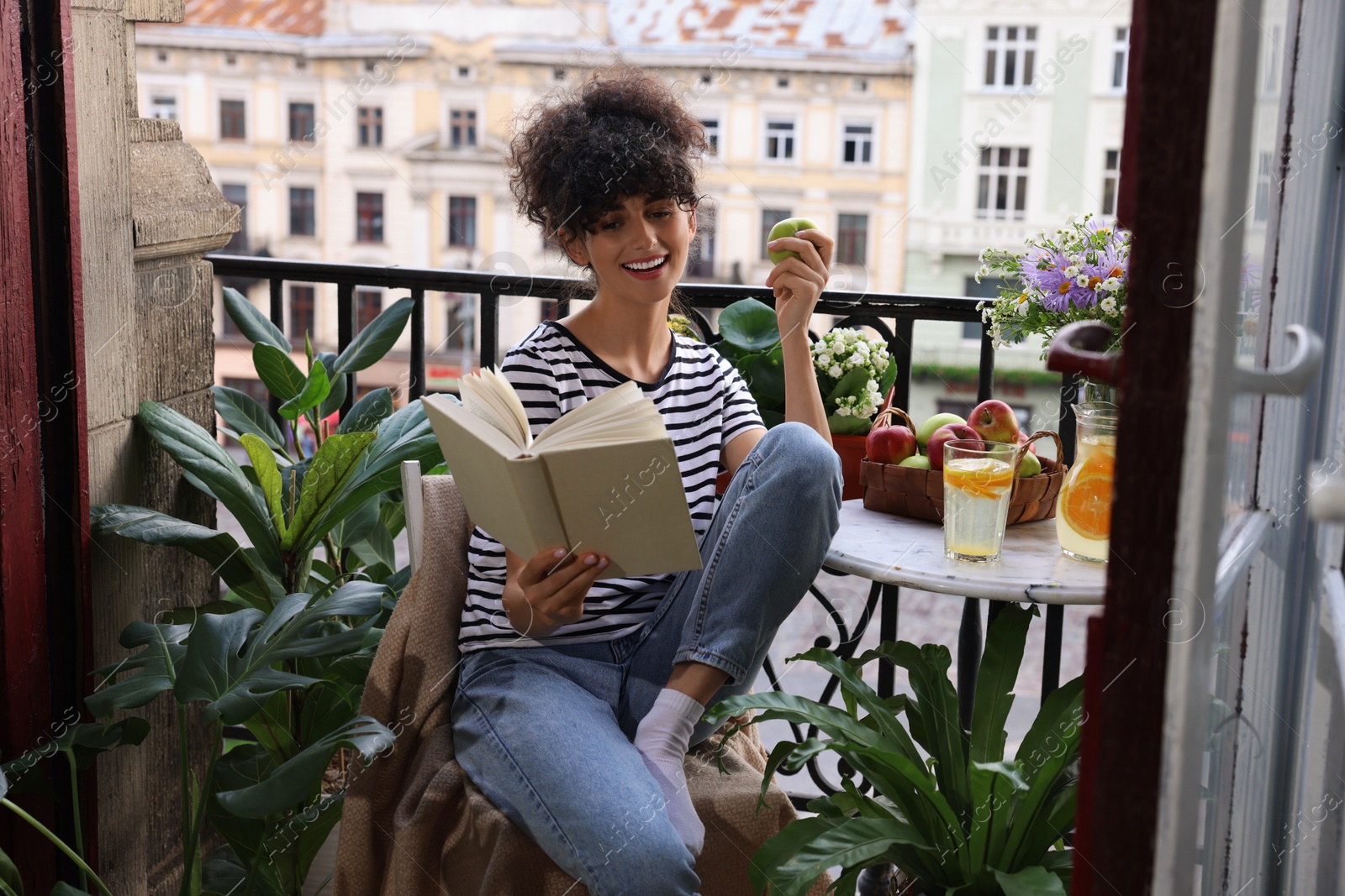 Photo of Young woman reading book at table on balcony with beautiful houseplants
