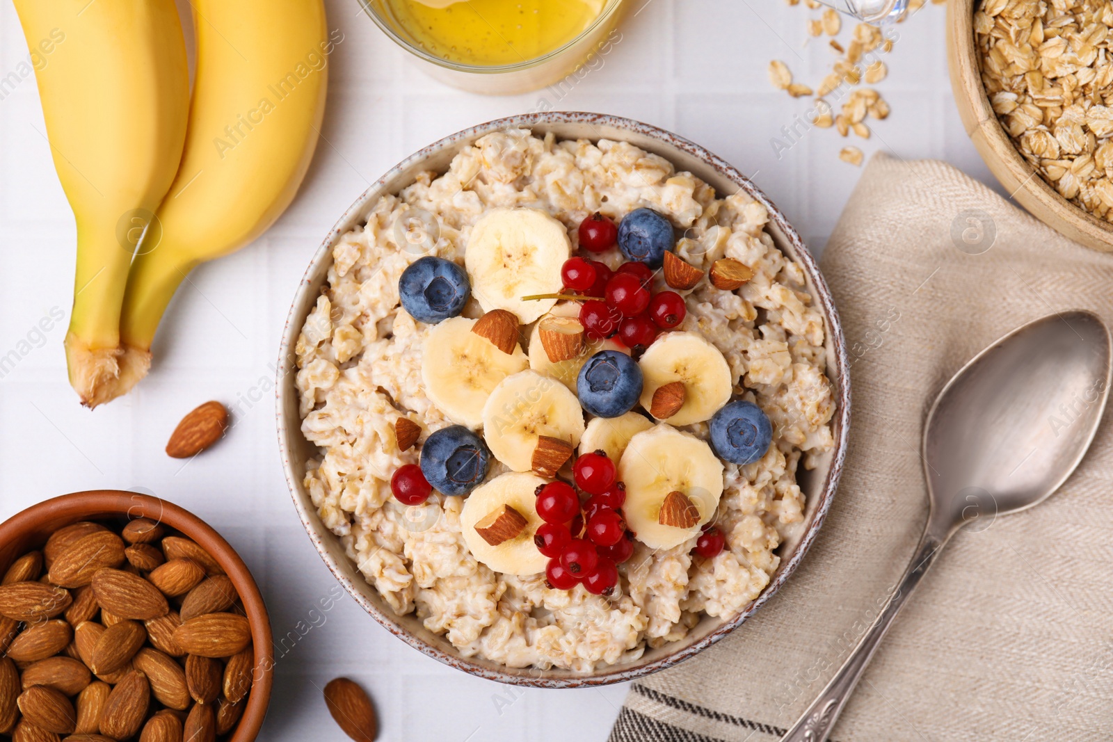 Photo of Oatmeal served with berries, almonds and banana slices on white tiled table, flat lay