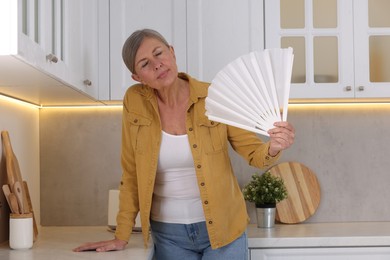 Photo of Menopause. Woman waving hand fan to cool herself during hot flash in kitchen