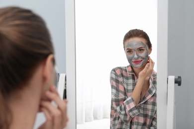 Photo of Young woman with cleansing mask on her face near mirror in bathroom. Skin care