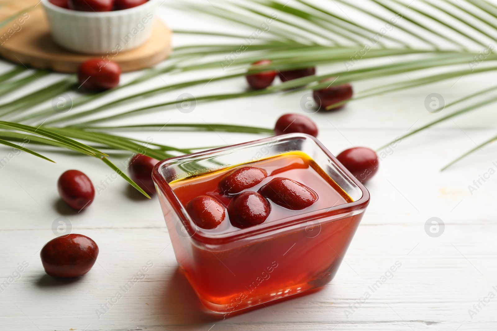 Photo of Palm oil in glass bowl with fruits and tropical leaves on white wooden table