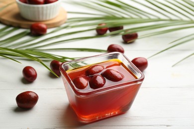 Photo of Palm oil in glass bowl with fruits and tropical leaves on white wooden table