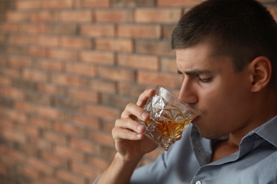 Young man with glass of whiskey near brick wall indoors. Space for text