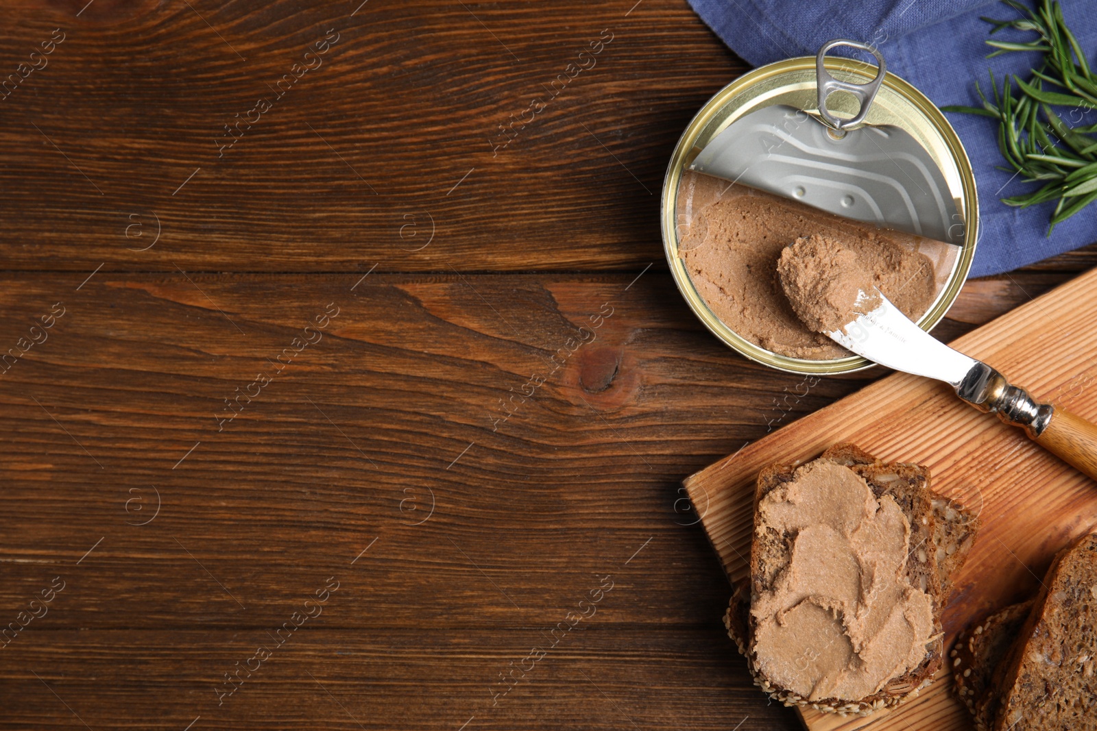 Photo of Open tin can with meat pate, fresh bread, knife and rosemary on wooden table, flat lay. Space for text