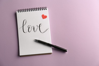 Photo of Notebook with handwritten word Love, red paper heart and pen on violet background, top view. Space for text