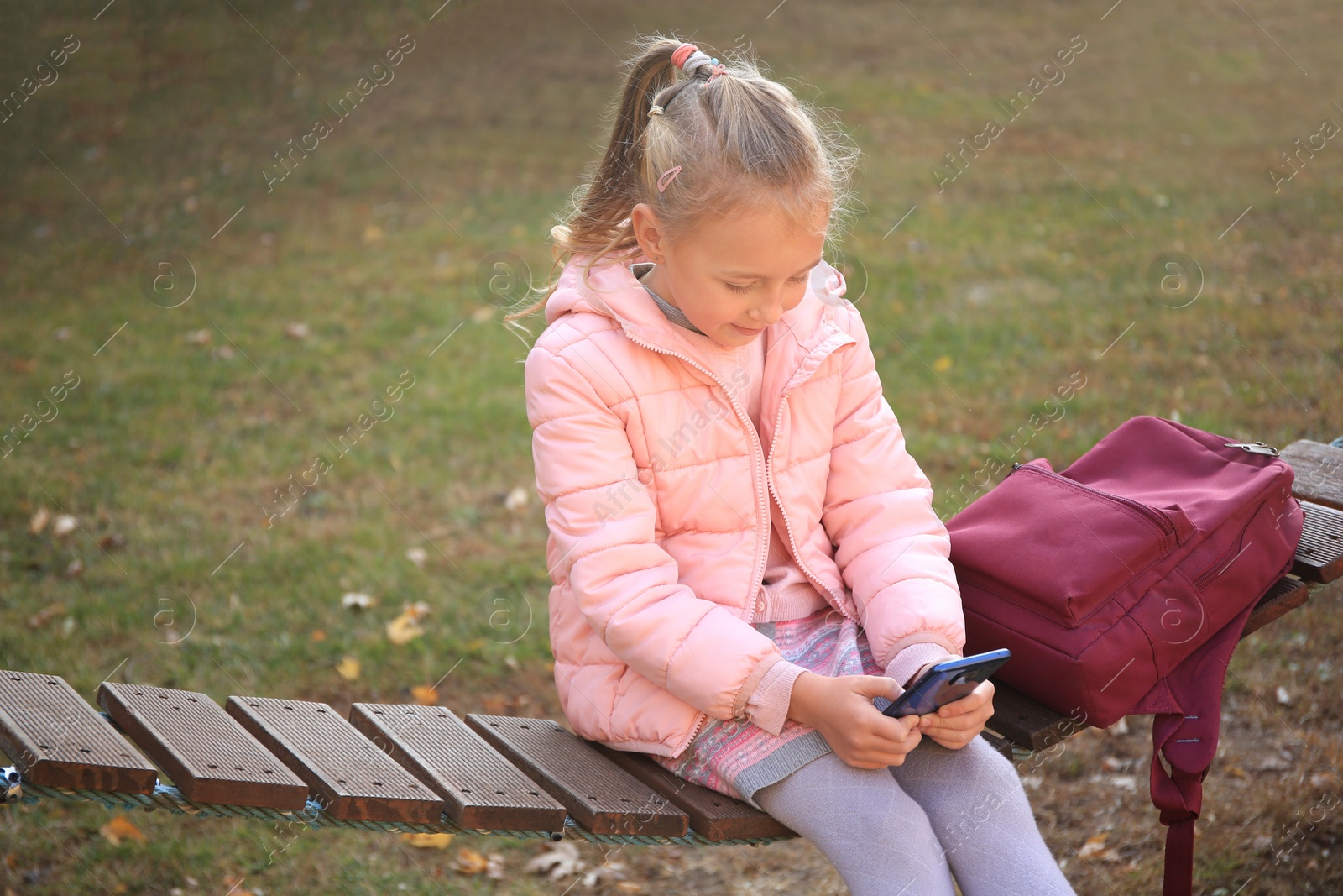 Photo of Cute little girl with backpack and smartphone on wooden swing outdoors