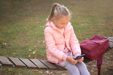 Photo of Cute little girl with backpack and smartphone on wooden swing outdoors