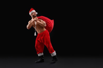 Photo of Muscular young man in Santa hat holding bag with presents on black background, space for text