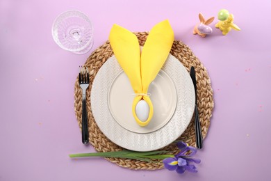 Festive table setting with painted egg, plates and iris flowers on lilac background, flat lay. Easter celebration