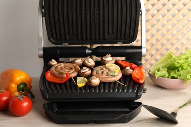 Photo of Electric grill with homemade sausages, vegetables and spatula on wooden table