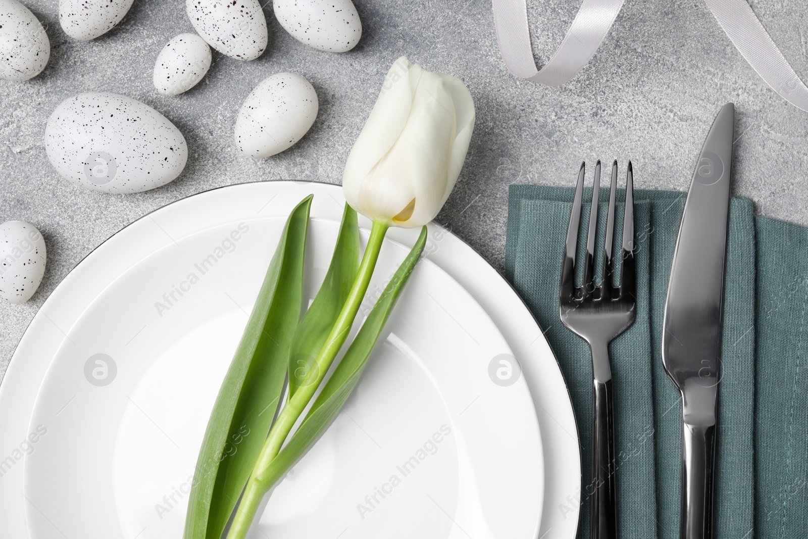 Photo of Festive table setting with painted eggs and white tulip on light grey background, flat lay. Easter celebration