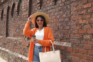 Young woman with stylish bag and cup of hot drink near red brick wall outdoors