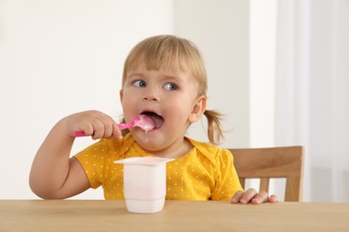 Cute little child eating tasty yogurt with spoon at wooden table indoors