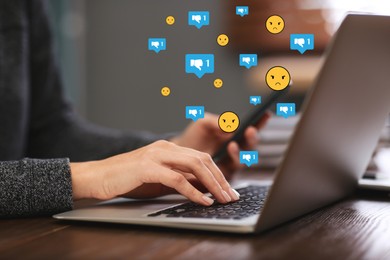Image of Social media dislike reaction. Woman using laptop and mobile phone at table, closeup. Thumbs down and angry face emoji illustrations over device