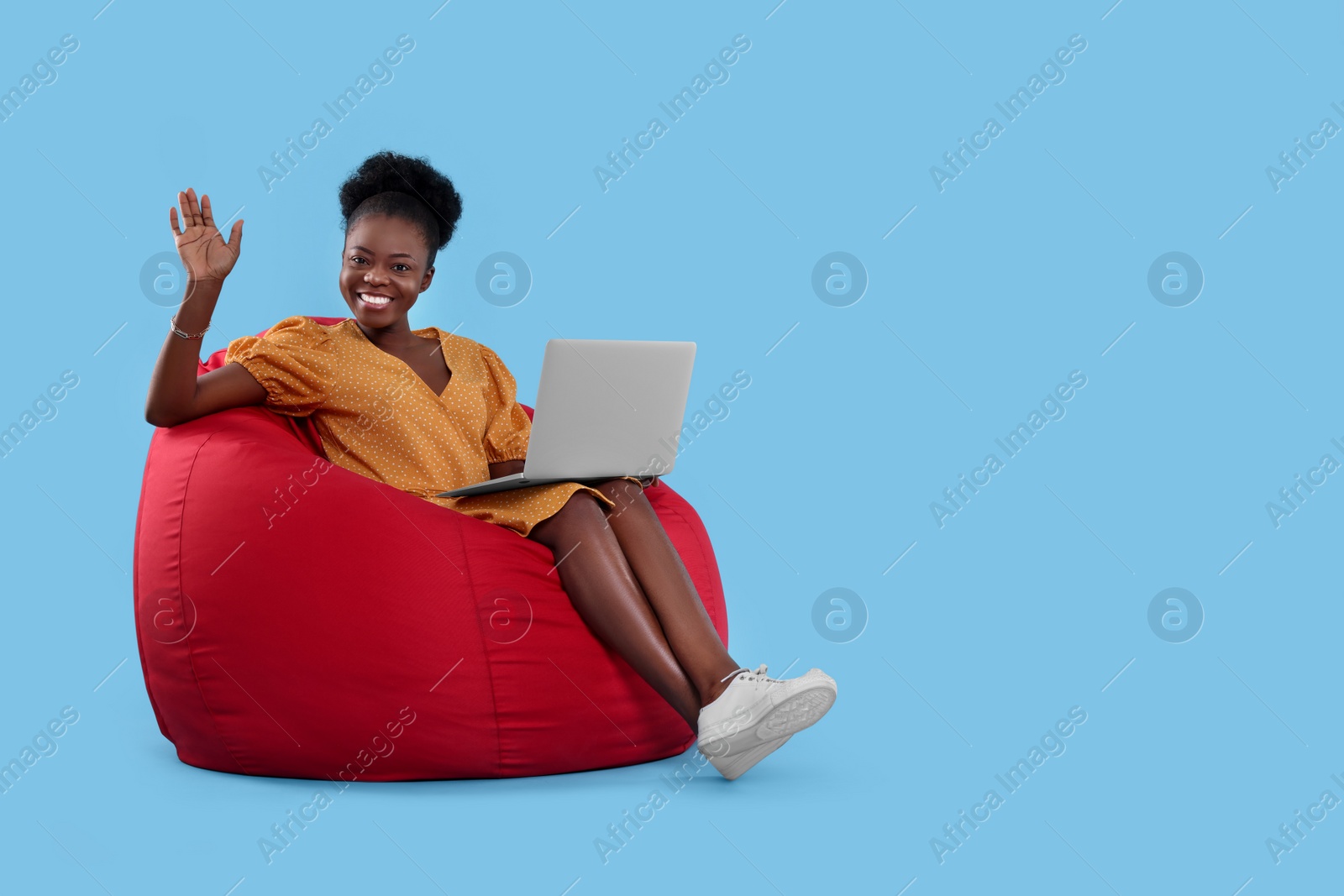 Photo of Beautiful young woman with laptop sitting on beanbag chair against light blue background. Space for text