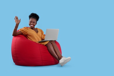 Photo of Beautiful young woman with laptop sitting on beanbag chair against light blue background. Space for text