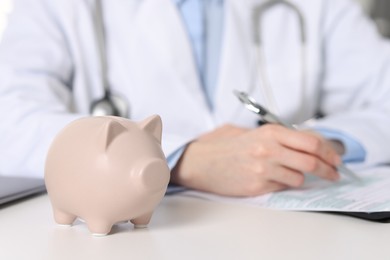 Photo of Doctor making notes at white table, focus on piggy bank