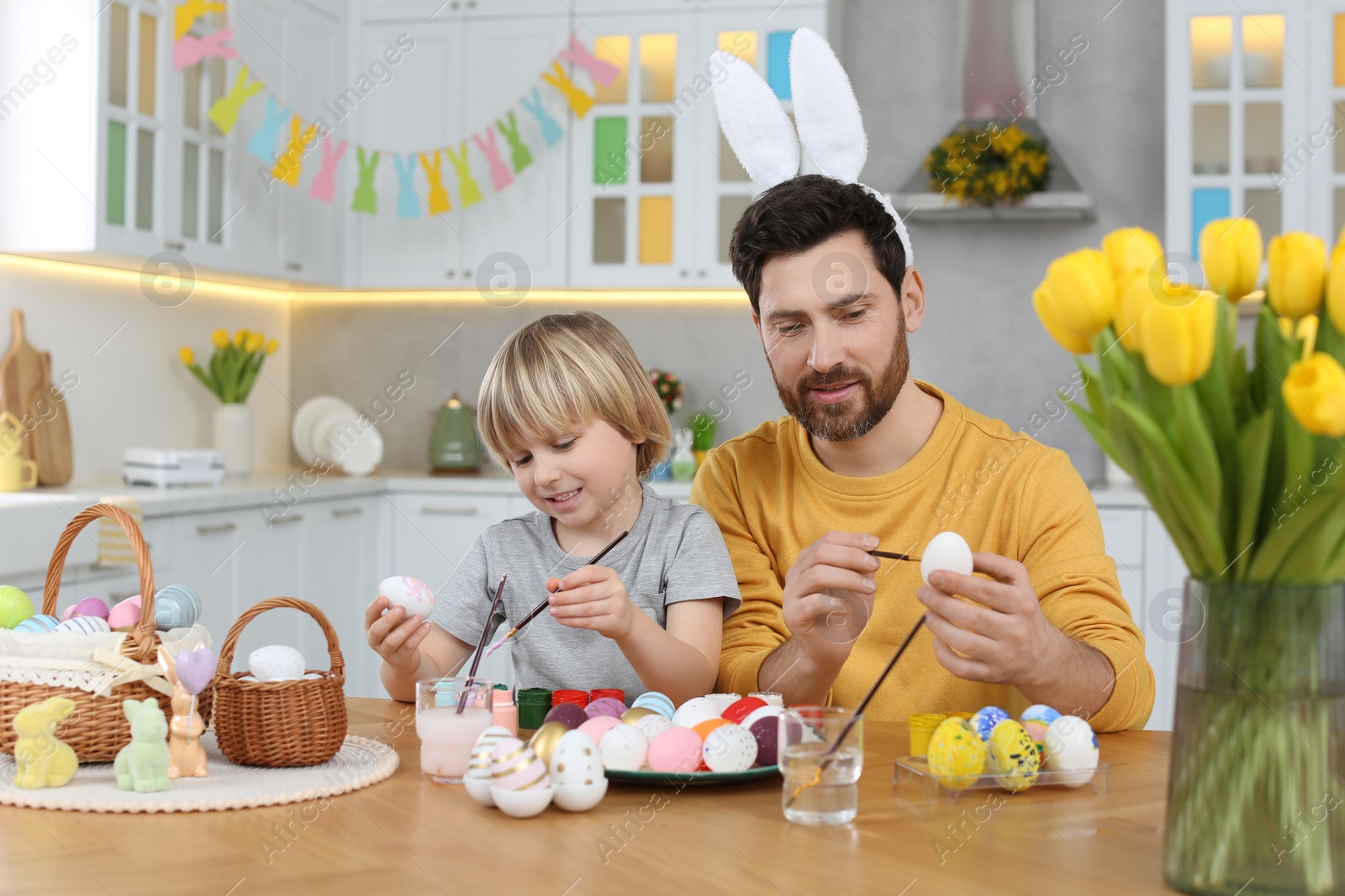 Photo of Father and his cute son painting Easter eggs at table in kitchen