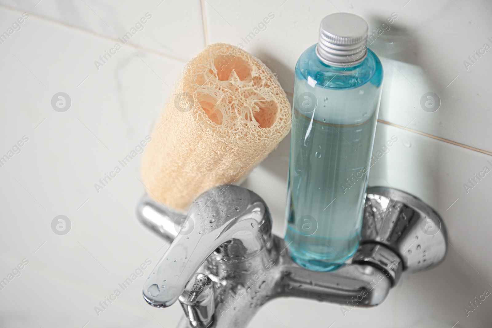 Photo of Natural loofah sponge and shower gel bottle on faucet in bathroom, closeup
