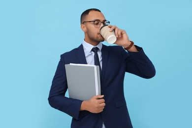 Young intern with notebooks drinking hot coffee on light blue background
