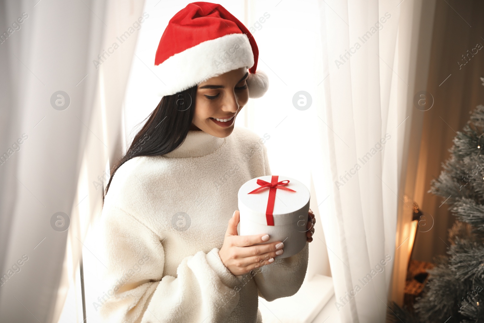 Photo of Woman in Santa hat holding gift box near Christmas tree