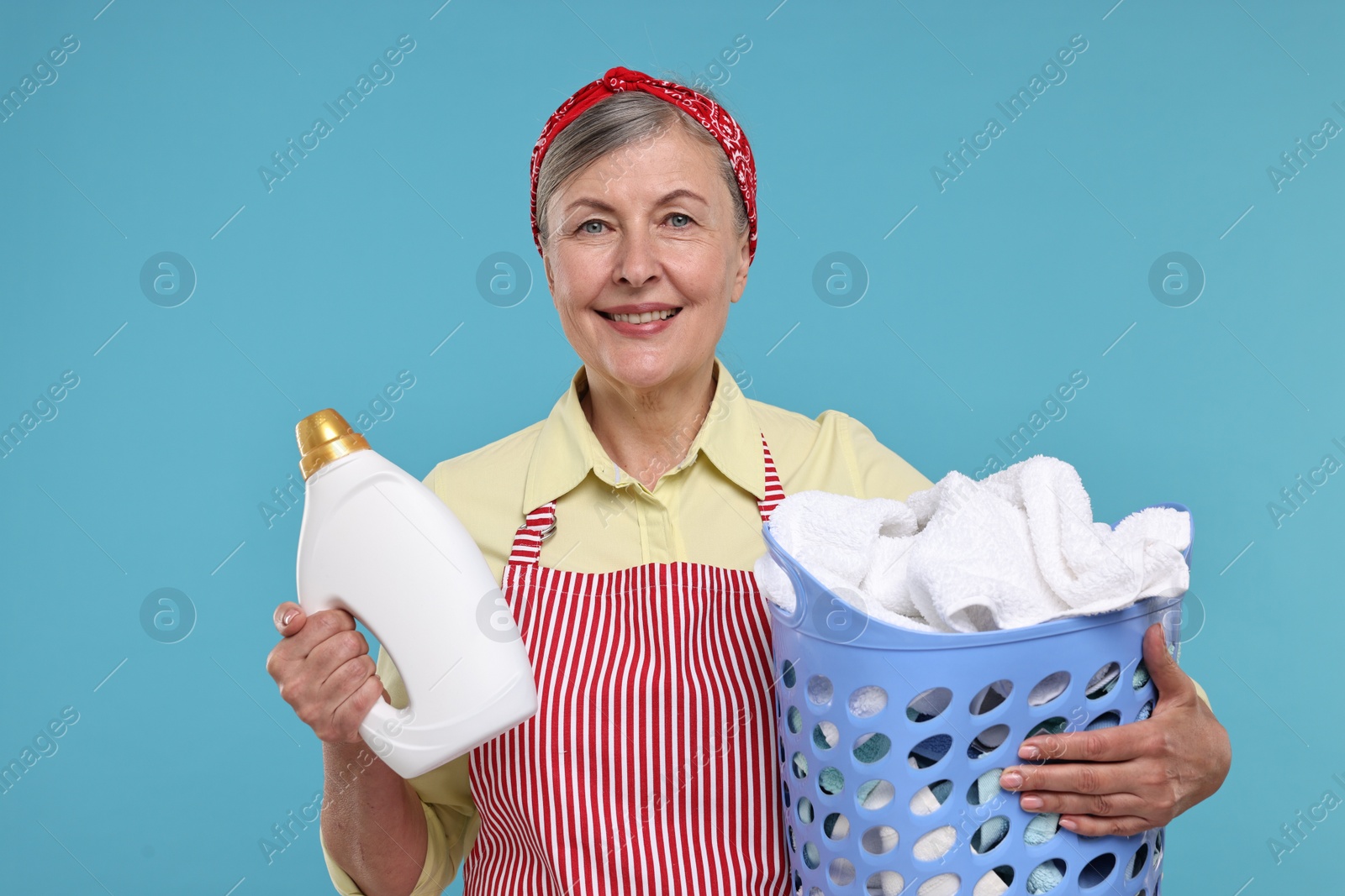 Photo of Happy housewife with detergent and basket full of laundry on light blue background