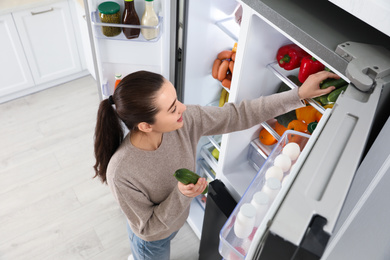 Photo of Young woman taking cucumber out of refrigerator indoors, above view