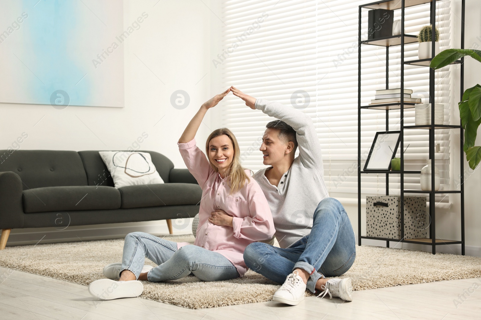 Photo of Young family housing concept. Pregnant woman with her husband forming roof with their hands while sitting on floor at home