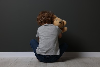 Photo of Child abuse. Upset boy with toy sitting on floor near grey wall, back view