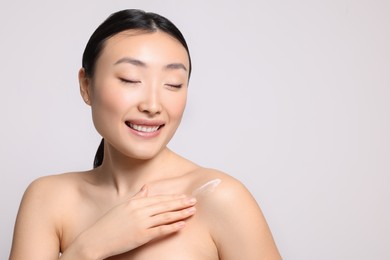 Photo of Beautiful young Asian woman applying body cream onto shoulder on light grey background, space for text